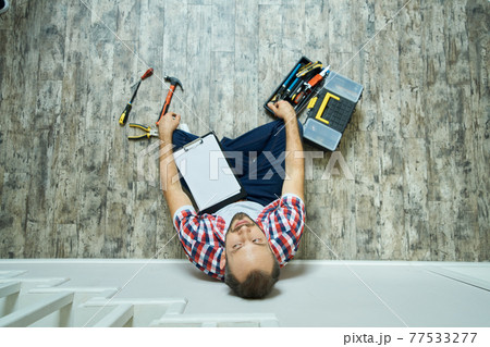 Top view of tired handyman taking a break, sitting with a clipboard on the floor. Home repair hand tools on laminate flooring 77533277