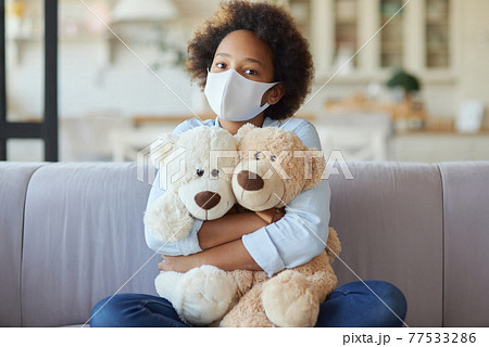 Portrait of cute mixed race teen girl in casual clothes wearing protective face mask, looking at camera, hugging her toys while sitting on a sofa at home 77533286