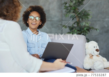 Cute teen girl smiling, listening to her female teacher, using laptop for studying while having a lesson, sitting on the couch at home 77533287