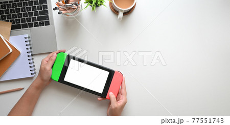 Top view image of hands holding a portable video game with white blank screen over white working desk. Top view image of hands holding a portable video game with white blank screen over white working desk. 77551743