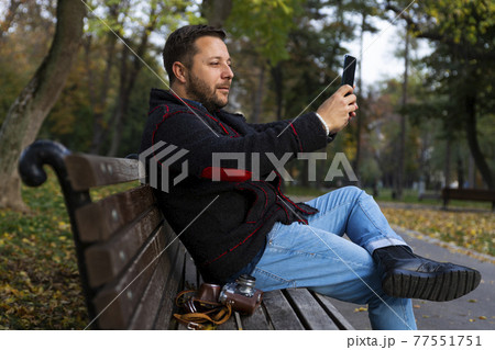 Man with smartphone in his hand taking selfie while sitting on the bench in the park at autumn 77551751