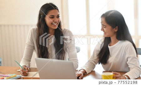 Young university student tutoring their lesson with using a computer laptop while sitting together at wood working desk over comfortable sitting room windows as background. Young university student tutoring their lesson with using a computer laptop while sitting together at wood working desk over comfortable sitting room windows as background. 77552198