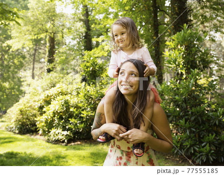 Mother giving toddler daughter a shoulder carry in park 77555185