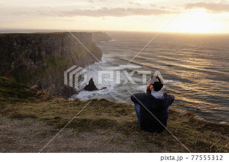 Mid adult man sitting on The Cliffs of Moher, The Burren, County Clare, Ireland 77555312