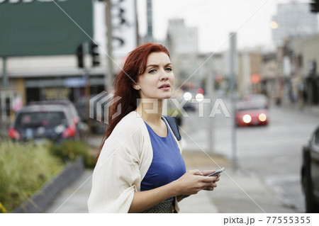 Young woman with long red hair, using smartphone in street 77555355