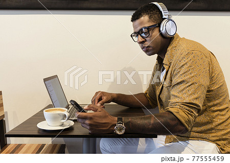 Young man in coffee shop, wearing headphones, using laptop and smartphone 77555459