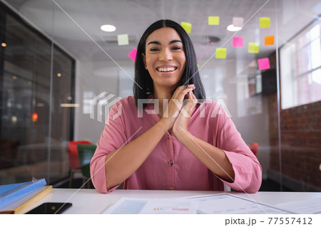 Happy mixed race businesswoman sitting at desk while having a video call smiling Happy mixed race businesswoman sitting at desk while having a video call smiling 77557412