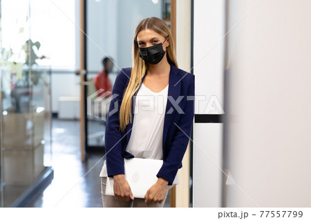 Caucasian businesswoman wearing mask standing in corridor holding laptop looking to camera Caucasian businesswoman wearing mask standing in corridor holding laptop looking to camera 77557799