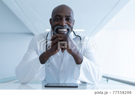 Smiling african american male doctor sitting at desk with tablet giving video call consultation 77558206