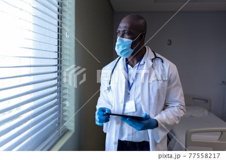 African american male doctor in hospital wearing face mask using tablet 77558217