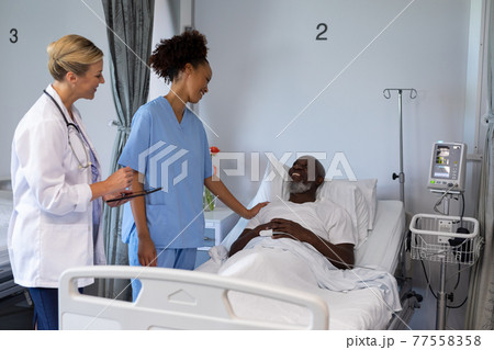 Two diverse female doctors and african american male patient in hospital room smiling to each other 77558358