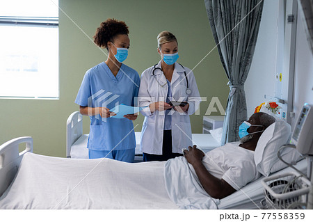 Two diverse female doctors wearing face masks talking with african american male patient 77558359