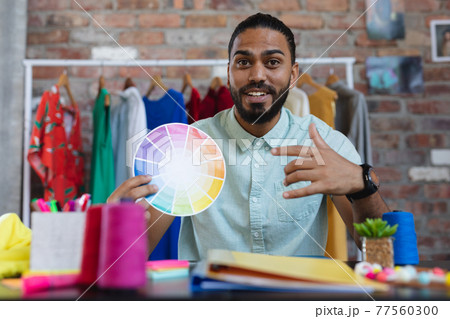 Mixed race male designer in sitting at desk holding a colour chart and smiling 77560300