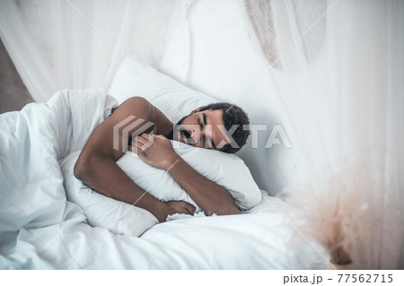 Dark-skinned man sleeping hugging pillow in bedroom Dark-skinned man sleeping hugging pillow in bedroom 77562715