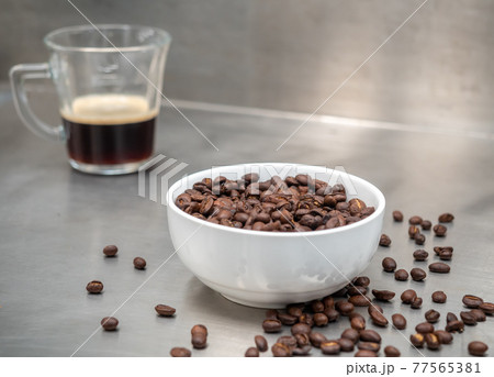 Roasted coffee beans in white bowl with americano in glass cup on table Roasted coffee beans in white bowl with americano in glass cup on table 77565381