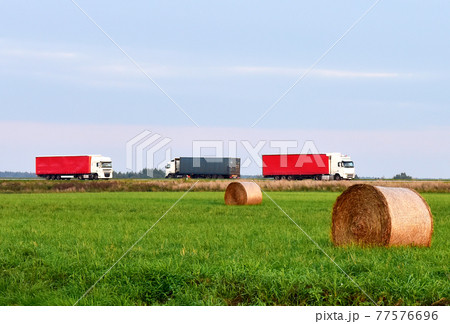 View of a field with hay in rolls against the background of trucks with semi-trailer driving View of a field with hay in rolls against the background of trucks with semi-trailer driving 77576696