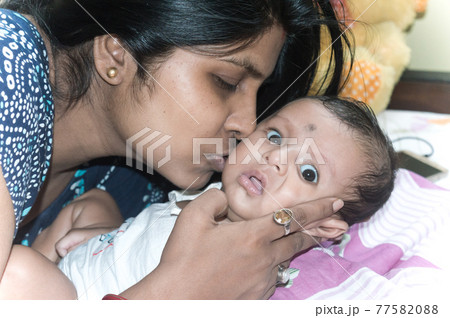 Close up Portrait of cute baby boy being kissed by mother on bed. Moms Love and mother's day background. Sweet child little infant toddler being hugged by mum. Front view. Close up Portrait of cute baby boy being kissed by mother on bed. Moms Love and mother's day background. Sweet child little infant toddler being hugged by mum. Front view. 77582088