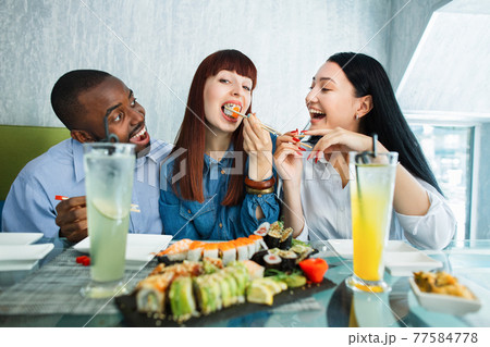 Sushi concept, seafood, asian meal. Three young multiethnic friends, Caucasian and Asian girls, African man, sitting in restaurant, spending funny time with plate of delicious sushi. 77584778