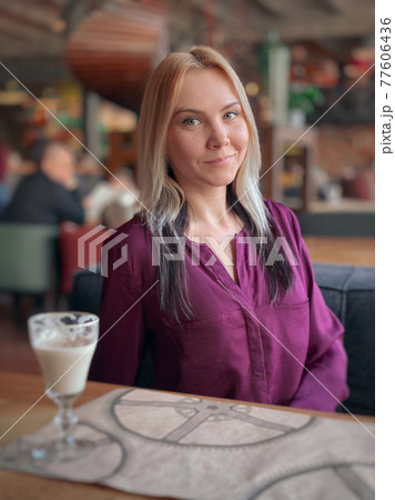 A blonde girl in a lilac sweater sits in a cozy restaurant on a sofa and she is relaxed and smiling and have a coffee drink on her table. 77606436
