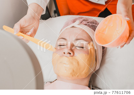 Woman at the beautician applying a relaxing golden mask on the face, restoring and moisturizing the skin. Woman at the beautician applying a relaxing golden mask on the face, restoring and moisturizing the skin. 77611706