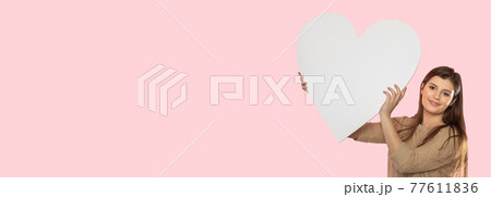 Girl with a large white heart shape in the hands. A student in love stands and holds a large heart shape in her hands. Valentine's Day and love. Panoramic frame. 77611836