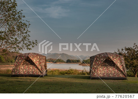 View of two camping tent with awning pitched on the dry ground Near beautiful big trees with branches at the campsite in lake shore with mountain range in background. View of two camping tent with awning pitched on the dry ground Near beautiful big trees with branches at the campsite in lake shore with mountain range in background. 77620567