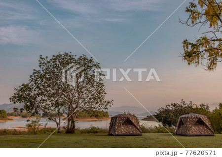 View of two camping tent with awning pitched on the dry ground Near beautiful big trees with branches at the campsite in lake shore with mountain range in background. View of two camping tent with awning pitched on the dry ground Near beautiful big trees with branches at the campsite in lake shore with mountain range in background. 77620571