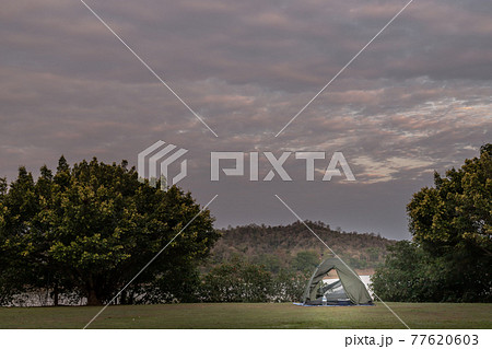 View of a camping tent with awning pitched on the dry ground near beautiful big trees with branches at the campsite lake shore with mountain range in background. View of a camping tent with awning pitched on the dry ground near beautiful big trees with branches at the campsite lake shore with mountain range in background. 77620603