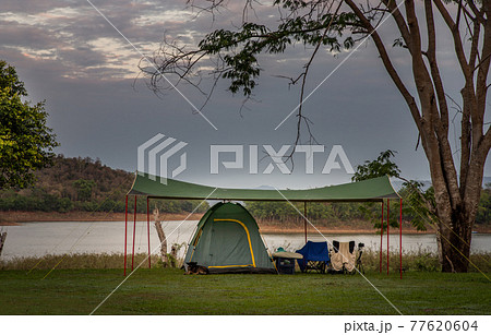 View of a camping tent with awning pitched On the grass under beautiful big trees with branches at the campsite lake shore with mountain range in background. View of a camping tent with awning pitched On the grass under beautiful big trees with branches at the campsite lake shore with mountain range in background. 77620604