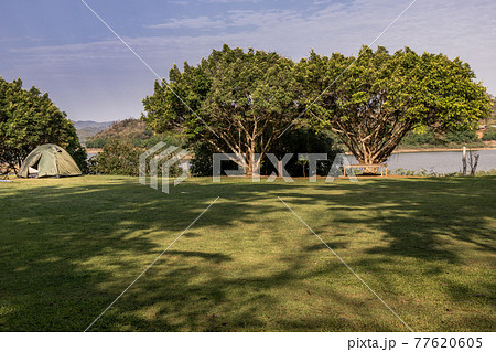 View of a camping tent with awning pitched on the dry ground near beautiful big trees with branches at the campsite lake shore with mountain range in background. View of a camping tent with awning pitched on the dry ground near beautiful big trees with branches at the campsite lake shore with mountain range in background. 77620605