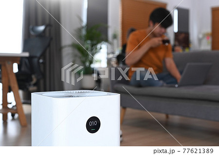 Air purifier on the floor in living room and young man sitting on sofa in background. Air purifier on the floor in living room and young man sitting on sofa in background. 77621389