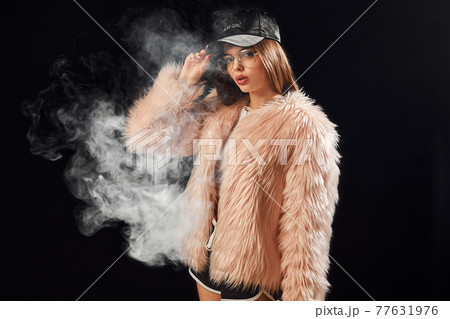 Sexy trendy rap woman in pink fur coat and cap smoke electronic cigarette. Dark Studio background shooting Sexy trendy rap woman in pink fur coat and cap smoke electronic cigarette. Dark Studio background shooting 77631976
