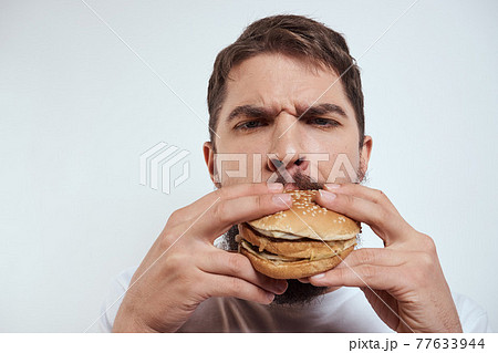 A man eating a hamburger on a light background in a white T-shirt cropped view close-up hunger fast food A man eating a hamburger on a light background in a white T-shirt cropped view close-up hunger fast food 77633944