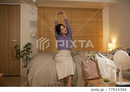 帰宅後ベッドに座り伸びをする女性 77634360