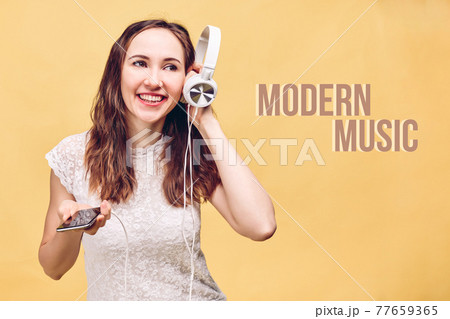 The girl on the yellow background smiles and listens to modern music through white headphones through her smartphone. Abstract inscription. The girl on the yellow background smiles and listens to modern music through white headphones through her smartphone. Abstract inscription. 77659365