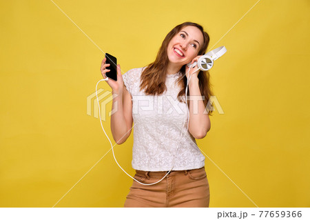 A girl in summer clothes stands on a yellow background and picks up music in a smartphone while listening to in the earphone. A girl in summer clothes stands on a yellow background and picks up music in a smartphone while listening to in the earphone. 77659366