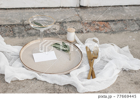 Festive wedding summer table setting. Golden cutlery, olive branch, glass of wine and silk ribbon on dinner plate. Grunge concrete background. Blank business card, invittation mockup. Top view. 77669445