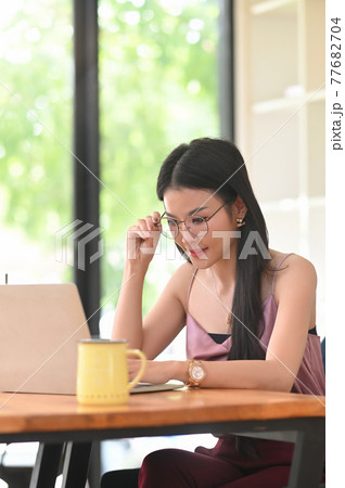 A woman with glasses is using a computer laptop while sitting at the wooden working desk. A woman with glasses is using a computer laptop while sitting at the wooden working desk. 77682704