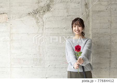 コンクリートの壁の前で薔薇を1輪持ち立っている若い女性 77684412
