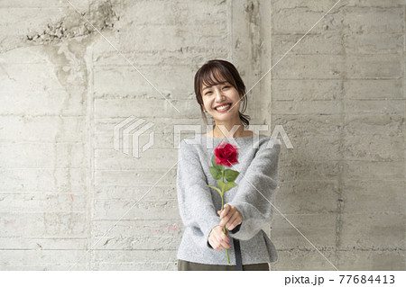 コンクリートの壁の前で薔薇を1輪持ち立っている若い女性 77684413
