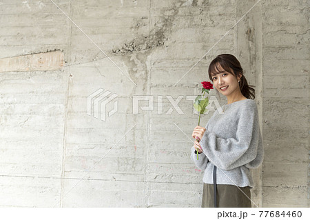 コンクリートの壁の前で薔薇を1輪持ち立っている若い女性 コンクリートの壁の前で薔薇を1輪持ち立っている若い女性 77684460