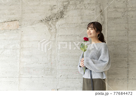 コンクリートの壁の前で薔薇を1輪持ち立っている若い女性 コンクリートの壁の前で薔薇を1輪持ち立っている若い女性 77684472