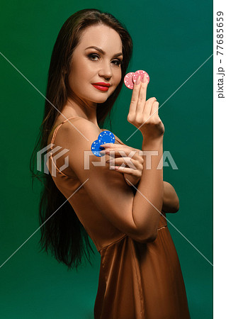 Brunette girl in silk beige dress. Smiling, showing four colorful chips, posing sideways on green background. Poker, casino. Close-up Brunette girl in silk beige dress. Smiling, showing four colorful chips, posing sideways on green background. Poker, casino. Close-up 77685659