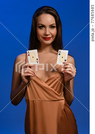 Brunette model in silk beige dress. She smiling, showing two aces, posing against blue background. Gambling, poker, casino. Close-up 77685685