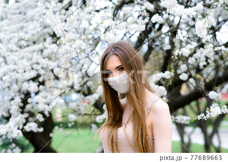 Close up portrait of tender girl in a white blouse under a blossoming cherry tree with a mask from the coronavirus. Close up portrait of tender girl in a white blouse under a blossoming cherry tree with a mask from the coronavirus. 77689663