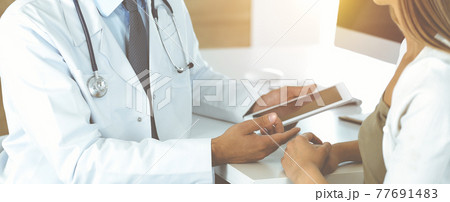 Doctor and patient discussing medical exam results while sitting at the desk in sunny clinic, close-up. Male physician using tablet computer for filling up medication history record. 77691483