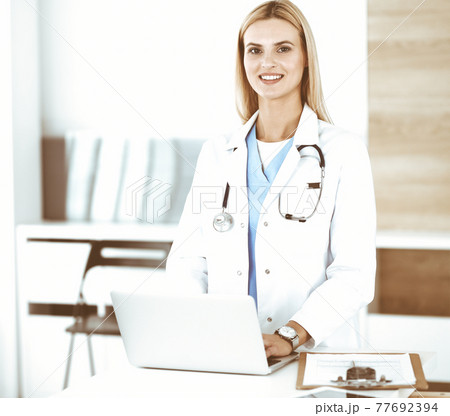 Blond female physician is lsmiling while using laptop computer. Woman-doctor at work in clinic excited and happy of her profession. Medicine concept Blond female physician is lsmiling while using laptop computer. Woman-doctor at work in clinic excited and happy of her profession. Medicine concept 77692394