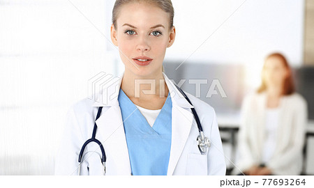 Woman - doctor is using clipboard for filling up medication history record form in sunny clinic at the background of patient 77693264