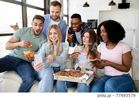 Positive diverse friends having home party, sitting on couch with pizza, chips and smartphone, having fun together 77693579
