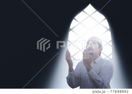 Asian Muslim man sitting while raised hands and praying inside the room Asian Muslim man sitting while raised hands and praying inside the room 77698892